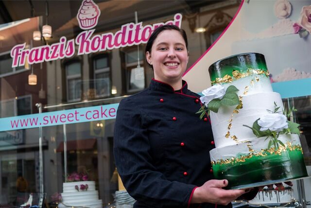 Eine Frau in Konditoren-Kleidung hält eine Torte vor der Glasfront eines Ladenschäfts mit der Aufschrift „Franzis Konditorei“.