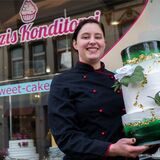 Eine Frau in Konditoren-Kleidung hält eine Torte vor der Glasfront eines Ladenschäfts mit der Aufschrift „Franzis Konditorei“.