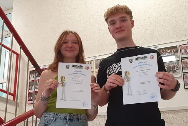 Ein Junge und ein Mädchen stehen im Flur einer Schule und halten Urkunden in den Händen.