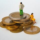 Eine Frau und ein Mann im Miniaturformat sitzen beziehungswiesen stehen auf Eurostücken. Der Mann steht über der Frau. Beide stehen sich nicht an.