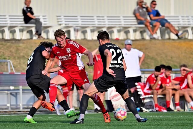 Zwei Spieler vom TuS Haltern im Dreikampf mit einem Spieler vom VfB Hüls; im Hintergrund sind Auswechselbank und Trainer des VfB Hüls unscharf sichtbar.