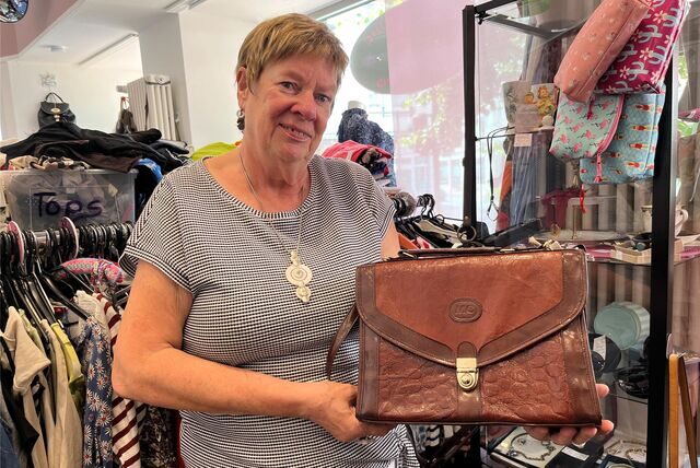 Heike Neumann hält in ihrem Laden der Castroper Altstadt eine braune Ledertasche in die Kamera. Im Hintergrund sieht man eine Vitrine mit Schmuck und voll bepackte Kleiderständer.