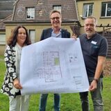 Drei Personen halten einen Bauplan hoch.