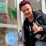 Yvonne Bobb neben dem Refill-Logo im Schaufenster. Es weist auf den kostenlosen Trinkwasser-Service des Friseursalons „Haar Zwei Ooh“ hin.