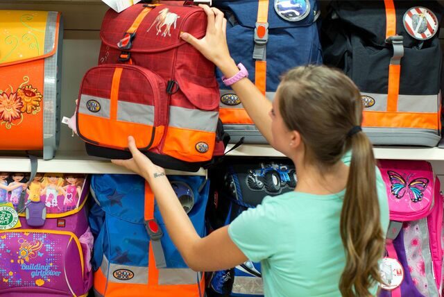 Eine Frau steht vor einem Regal, in dem verschiedene Tornister ausgestellt sind. Sie zieht einen roten Rucksack aus dem Regal und ist dabei von hinten zu sehen.