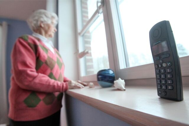 Eine Seniorin schaut aus dem Fenster. Auf der Fensterbank neben ihr steht ein Festnetz-Telefon. (Symbolbild)