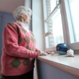 Eine Seniorin schaut aus dem Fenster. Auf der Fensterbank neben ihr steht ein Festnetz-Telefon. (Symbolbild)