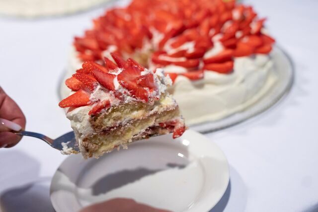 Ein Stück finnische Erdbeertorte auf einem Kuchenheber wird auf einen Teller gelegt. Im Hintergrund sieht man die restliche Torte auf einem Tisch stehen.