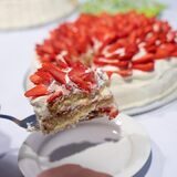 Ein Stück finnische Erdbeertorte auf einem Kuchenheber wird auf einen Teller gelegt. Im Hintergrund sieht man die restliche Torte auf einem Tisch stehen.