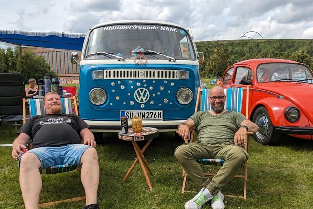 Ein historischer VW-Bus und ein Käfer stehen auf einer Wiese. Davor sitzen zwei Männer auf Klappstühlen.