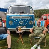 Ein historischer VW-Bus und ein Käfer stehen auf einer Wiese. Davor sitzen zwei Männer auf Klappstühlen.