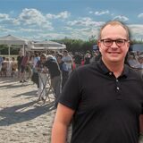 Eine Bildmontage: Im Hintergrund ist ein Bierwagen zu sehen, der auf hellem Strandsand steht. Drumherum stehen Partygäste an Tischen. Rechts im Bild ist der Sprecher der Bürgerschützengilde Datteln-Hagem, Duston Piotrowski, zu sehen.