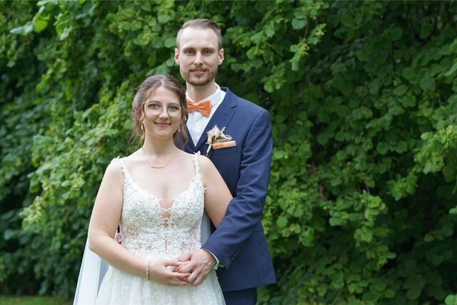 Rebecca und Gerrit stehen zusammen. Im Hintergrund sind grüne Bäume zu sehen.
