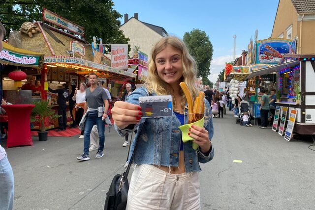 Eine junge Frau steht auf der Kirmes und hält ein DeutschlandTicket in die Kamera. In der Hand hält sie zudem einen Becher Churros.
