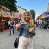 Eine junge Frau steht auf der Kirmes und hält ein DeutschlandTicket in die Kamera. In der Hand hält sie zudem einen Becher Churros.