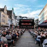 Auf dem Altstadtmarkt in Recklinghausen steht eine große Bühne, darauf spielt die Neue Philharmonie Westfalen. Auf Stühlen vor der Bühne sitzen Hunderte Zuhörer.