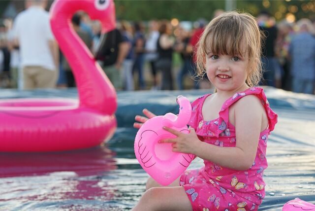 Ein kleines Mädchen sitzt am Rand eines Wasserbeckens und spielt mit einem pinkfarbenen Schwimmtier in Form eines Flamingos.