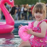 Ein kleines Mädchen sitzt am Rand eines Wasserbeckens und spielt mit einem pinkfarbenen Schwimmtier in Form eines Flamingos.