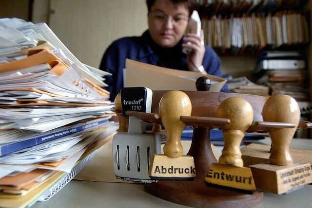 Eine Mitarbeiterin des Landratsamts Straubing-Bogen sitzt telefonierend hinter zahlreichen Akten und Stempeln an ihrem Arbeitsplatz.