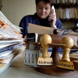 Eine Mitarbeiterin des Landratsamts Straubing-Bogen sitzt telefonierend hinter zahlreichen Akten und Stempeln an ihrem Arbeitsplatz.