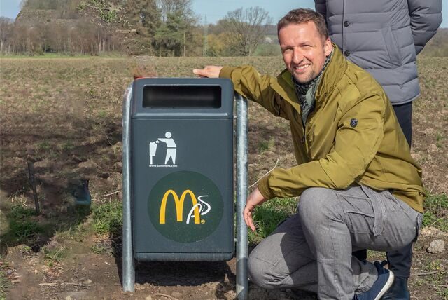 Markus von Gilardi hockt neben einem Mülleimer, auf dem ein McDonalds-Aufkleber zu sehen ist.