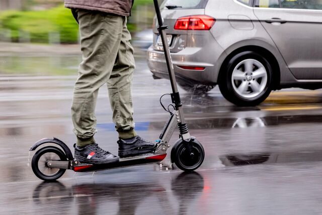 Ein Mann fährt mit einem E-Scooter auf einer Straße. Nur sein Unterkörper ist zu sehen. Im Hintergrund ein fahrendes Auto.