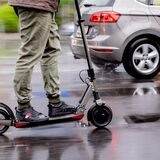 Ein Mann fährt mit einem E-Scooter auf einer Straße. Nur sein Unterkörper ist zu sehen. Im Hintergrund ein fahrendes Auto.
