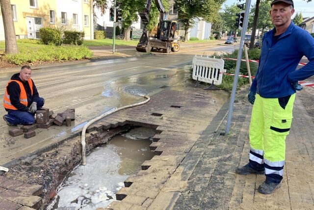 Zwei Männer in Arbeitskleidung stehen neben einem Loch in der Straße am Westring.