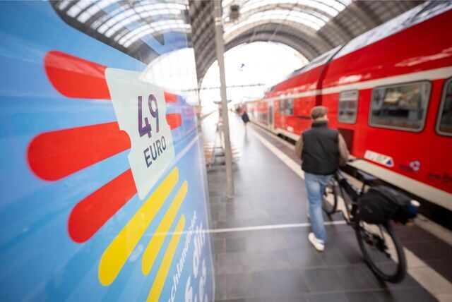 Ein Mann geht am Hauptbahnhof mit seinem Fahrrad zwischen der Werbung für das Deutschlandticket und einer Regionalbahn vorbei.