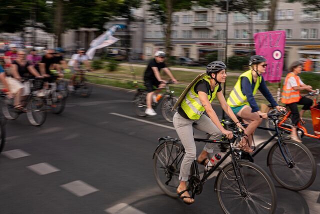 Fahrradfahrer sind zu sehen, wie sie auf einer Straße fahren. Sie tragen gelbe Warnwesten und werden von einer Kolonne begleitet.