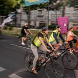 Fahrradfahrer sind zu sehen, wie sie auf einer Straße fahren. Sie tragen gelbe Warnwesten und werden von einer Kolonne begleitet.
