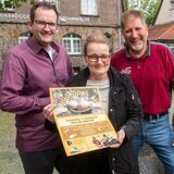 von links: Lukas Floßdorf (Stadt Datteln), Ulrike Köster („Lebendiger Tigg“) und Stephan Bröcker (Werbegemeinschaft) stehen mit einem Plakat an einem Brunnen und lächeln in die Kamera.