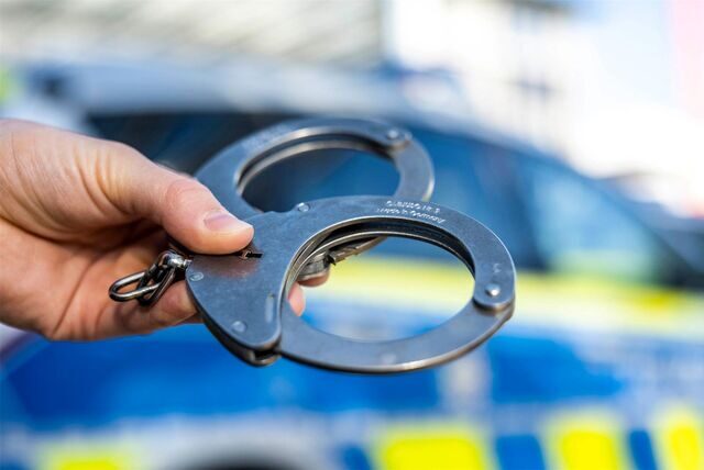 Eine Hand hält Handschellen vor einen Streifenwagen der Polizei (Symbolbild).