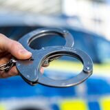 Eine Hand hält Handschellen vor einen Streifenwagen der Polizei (Symbolbild).