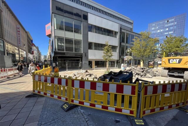 Gelbe Bauzäune umgeben die Baustelle in der Innenstadt. Trümmerteile und ein gelber Bagger sind zu sehen.