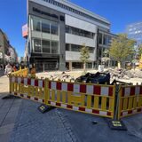 Gelbe Bauzäune umgeben die Baustelle in der Innenstadt. Trümmerteile und ein gelber Bagger sind zu sehen.
