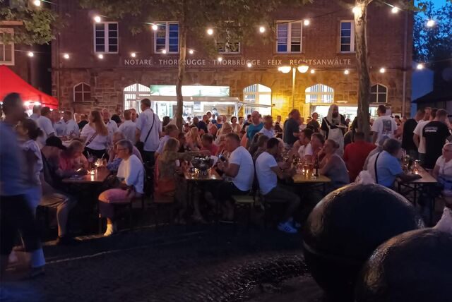 Menschen sitzen an Bierzeltgarnituren in der Dämmerung auf einem Platz.