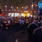 Menschen sitzen an Bierzeltgarnituren in der Dämmerung auf einem Platz.