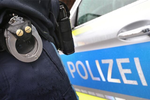 Der Gürtel eines uniformierten Polizisten mit Handschellen ist zu sehen, daneben die Tür eines Streifenwagens mit „Polizei“-Schriftzug.
