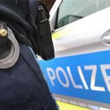 Der Gürtel eines uniformierten Polizisten mit Handschellen ist zu sehen, daneben die Tür eines Streifenwagens mit „Polizei“-Schriftzug.