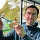 Auf der linken Seite liegt ein kaputtes Kameraobjektiv auf einem Tisch. Rechts ist der Besitzer dieses Objektives, Papatastisch-Autor Thomas Raulf.