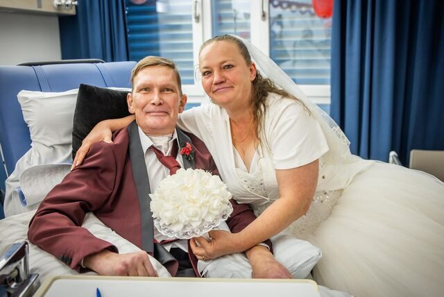 Das Ehepaar Waskönig im Krankenzimmer. Dirk sitzt in einem Stuhl und trägt einen Anzug. Die Braut umarmt ihn von der Seite.