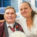 Das Ehepaar Waskönig im Krankenzimmer. Dirk sitzt in einem Stuhl und trägt einen Anzug. Die Braut umarmt ihn von der Seite.