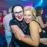 Zwei Frauen umarmen sich und schauen freudig in die Kamera. Im Hintergrund sind weitere Partybesucher zu sehen.