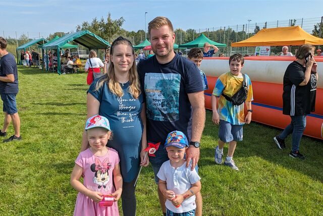 Eine Familie mit zwei Kindern lächelt in die Kamera. Im Hintergrund sind Stände und eine Hüpfburg zu sehen.