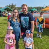 Eine Familie mit zwei Kindern lächelt in die Kamera. Im Hintergrund sind Stände und eine Hüpfburg zu sehen.