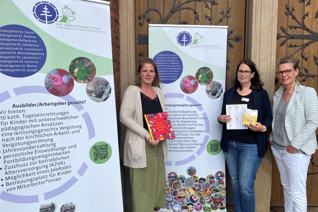 Drei Erzieherinnen mit großen Plakaten, die für die Ausbildung und Fachkräfte werben, vor der Kirchentür St. Sixtus