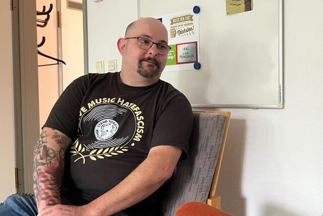 Ein Mann sitzt vor einem Whiteboard. Er spricht mit jemanden. Sein Arm ist tätowiert.