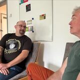 Ein Mann sitzt vor einem Whiteboard. Er spricht mit jemanden. Sein Arm ist tätowiert.