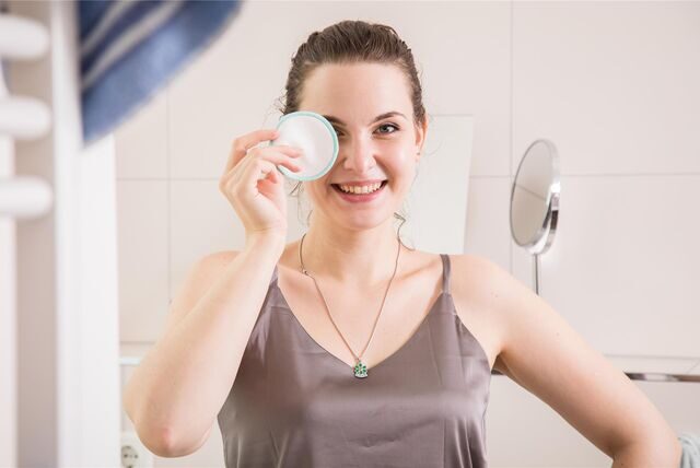 Eine Frau steht im Badezimmer. Sie lächelt in die Kamera und hält sich dabei ein wiederverwendbares Wattepad vor ein Auge.